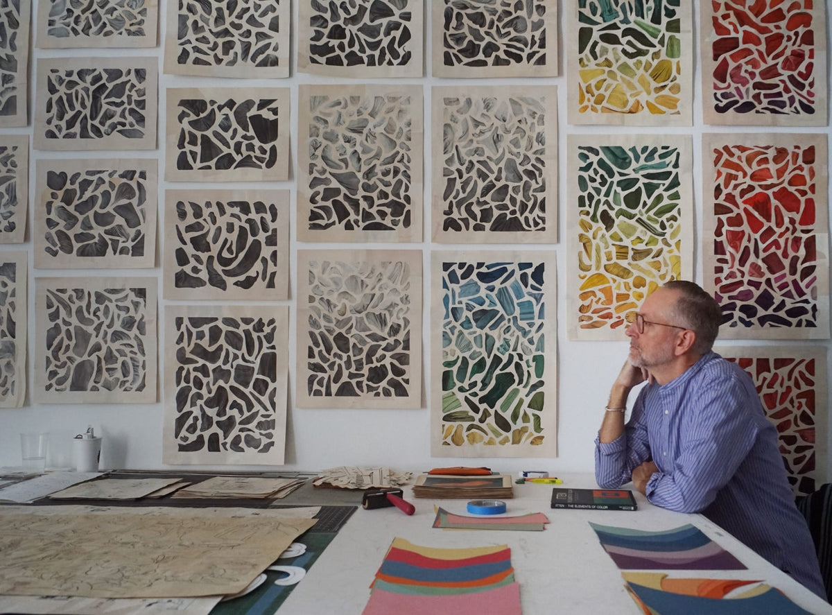 Artist in a blue button-down shirt working at a table covered with several framed intricate abstract line drawings