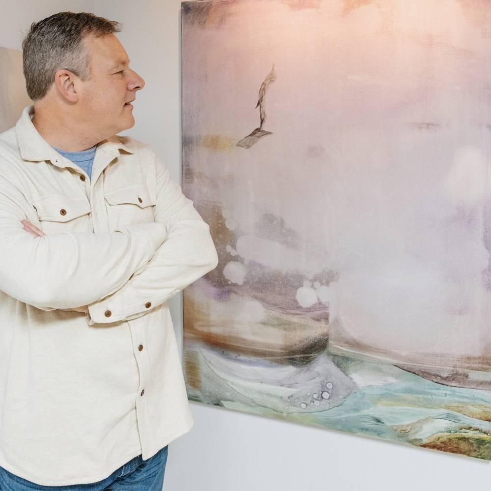A man in a cream button-down shirt standing with arms crossed beside a large ethereal pink and blue landscape painting