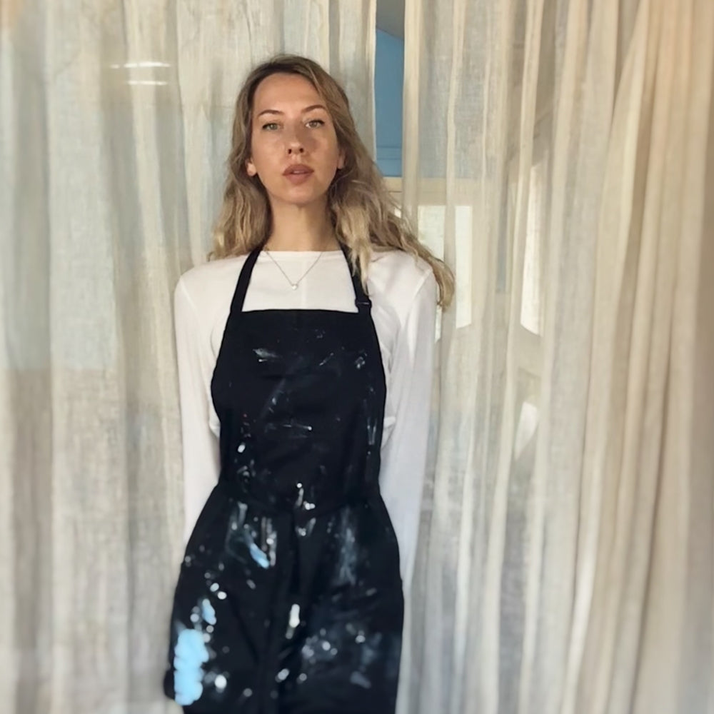 Woman with blonde wavy hair wearing a white long-sleeve shirt and a black apron splattered with white paint