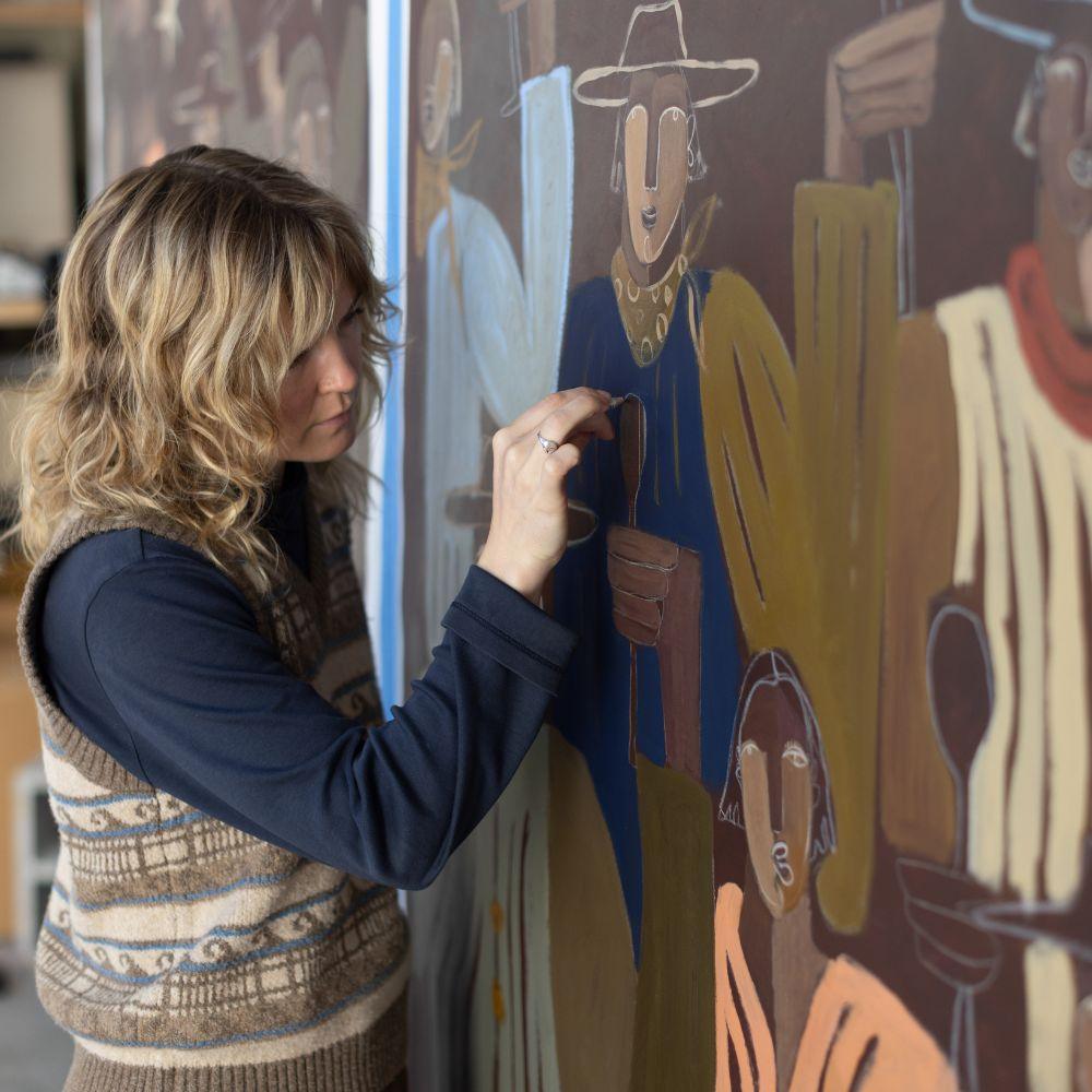 Artist in a brown knit vest and dark top painting a large-scale figurative mural with brown and colorful tones