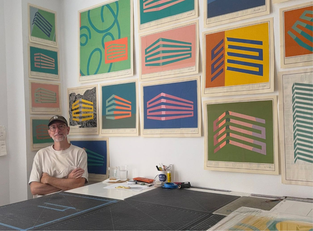 Artist in a studio next to a wall covered with colorful framed geometric art prints featuring building outlines