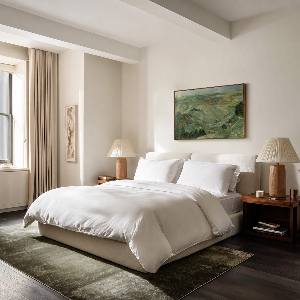 Country Hillside France painting in a bedroom hung above a bed with white linen and two wooden lamps