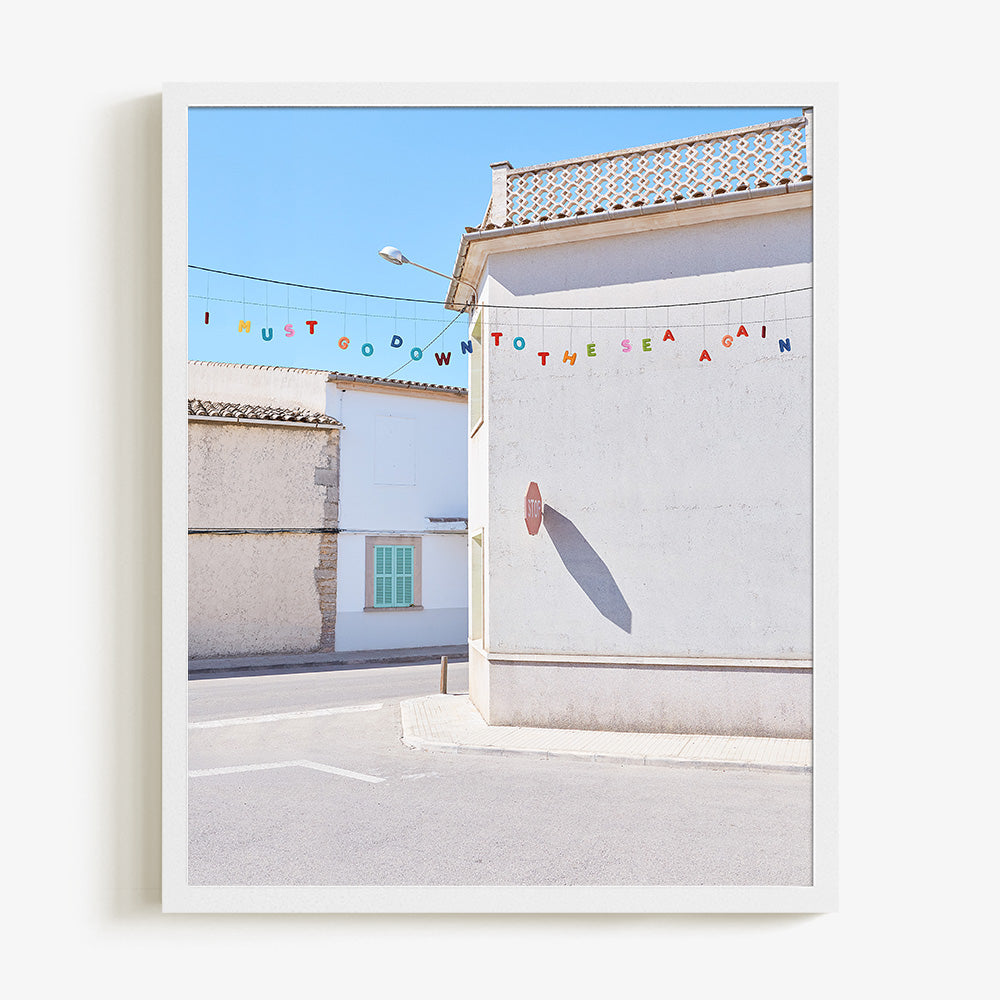 A white framed print of a white building with a stop sign shadow and colorful letters spelling I MUST GO DOWN TO THE