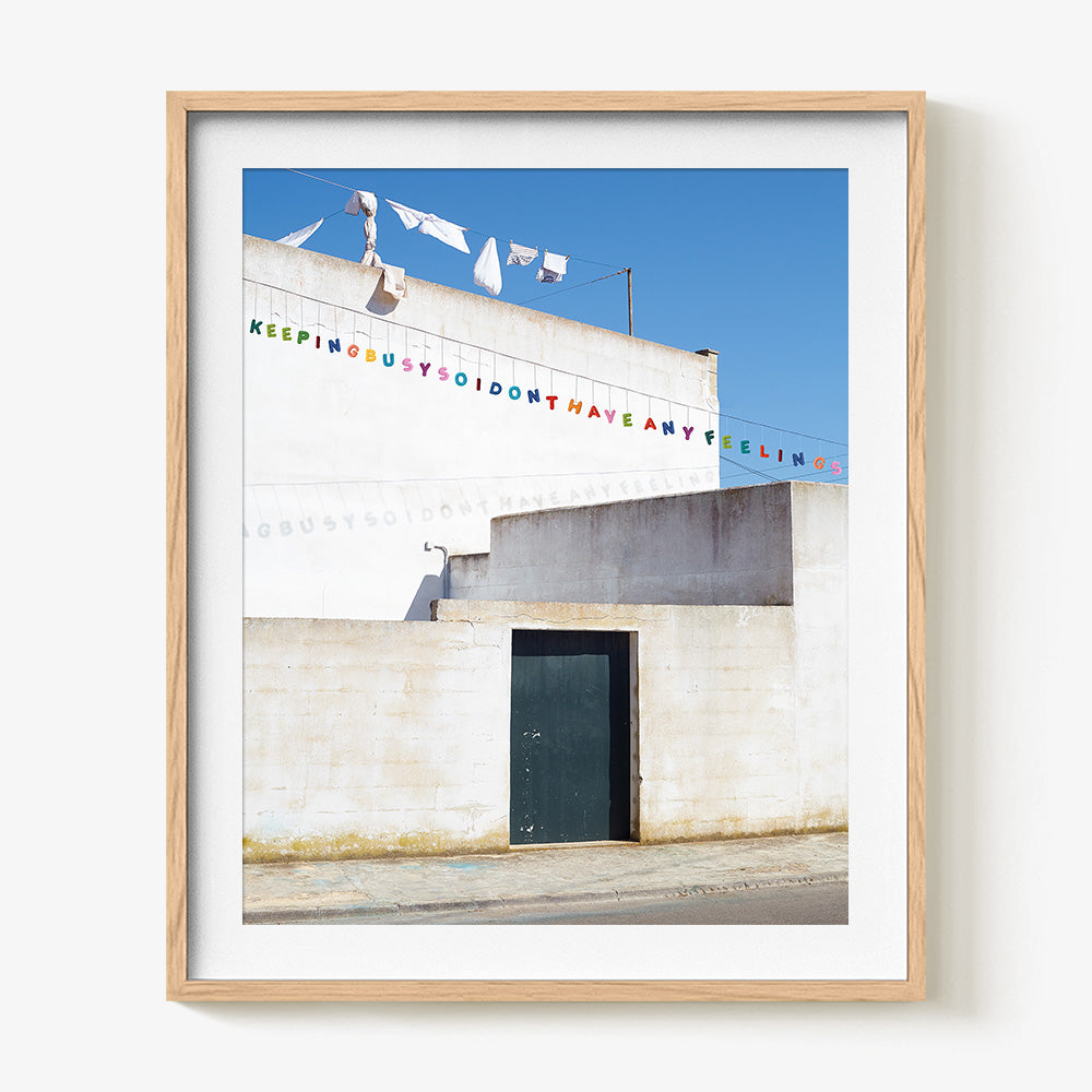 James Needham print of a white building with a dark door colorful banner text and laundry in a white frame