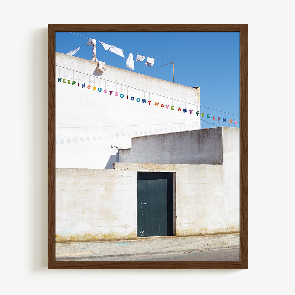 Print showing a white wall a dark green door and laundry hanging against a bright blue sky in a dark wood frame