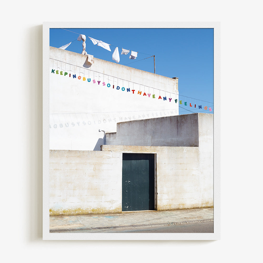 Print of a white wall with a dark door and colorful banner letters against a blue sky in a thin white frame