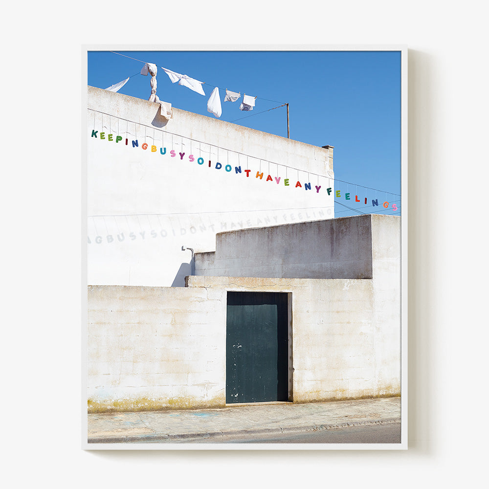 White building with laundry hanging and colorful letters spelling KEEPING BUSY SO I DONT HAVE ANY FEELINGS white frame