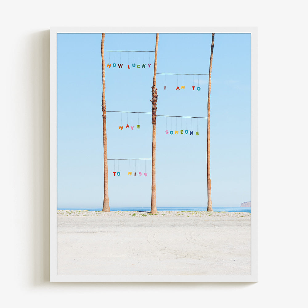 Lucky print by James Needham Colorful letters suspended between palm trees on a beach in a white frame