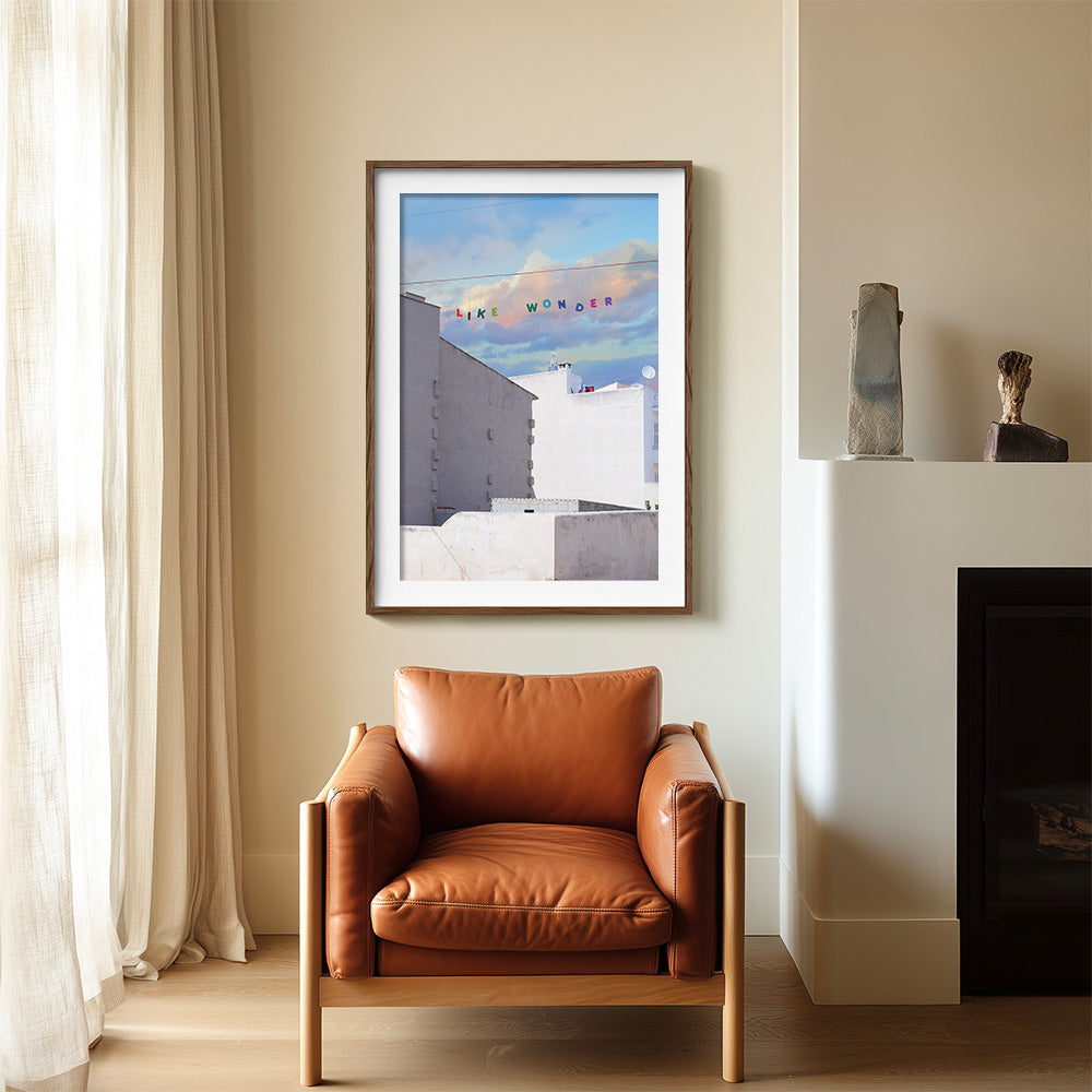 A print in a dark wood frame hanging above a brown leather armchair and fireplace depicting white buildings under a
