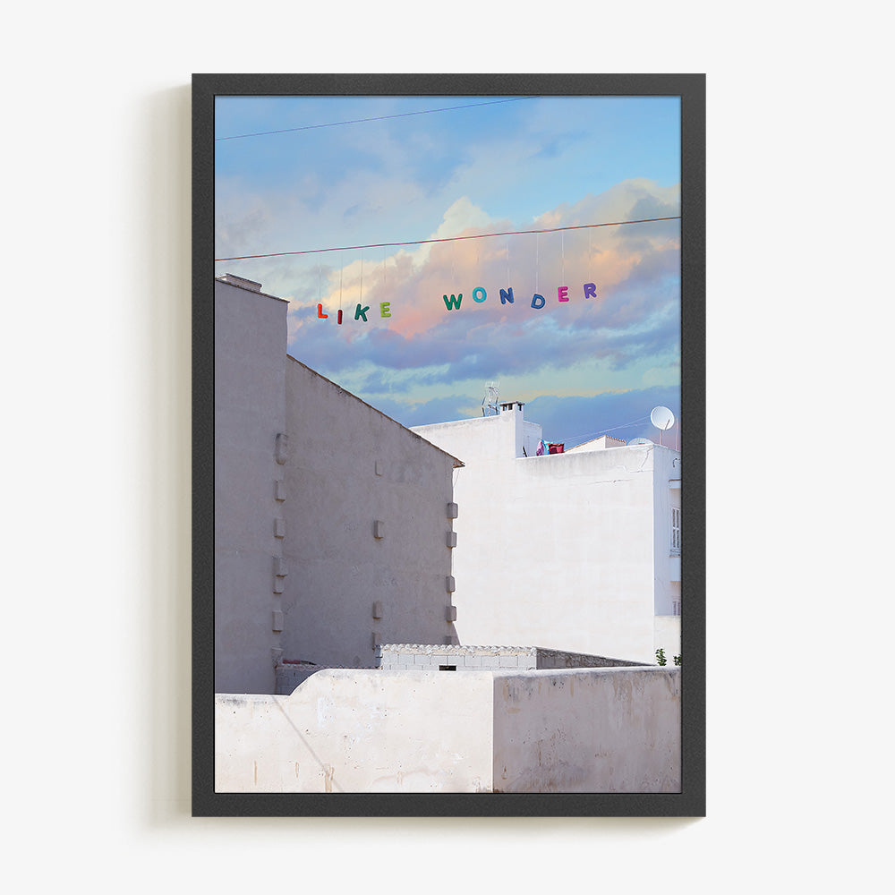 Art print of white buildings under a blue and purple cloudy sky with LIKE WONDER in colorful letters in a thin black