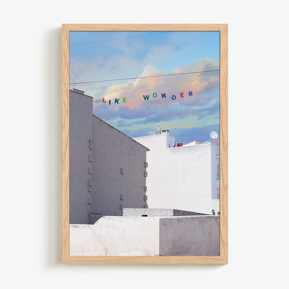 Art print of white buildings under a blue and purple cloudy sky with LIKE WONDER in colorful letters in a light wood