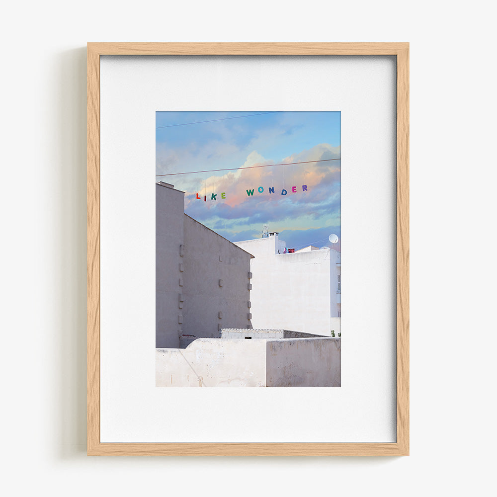 Art print of white buildings under a blue and purple cloudy sky with LIKE WONDER in colorful letters in a natural wood