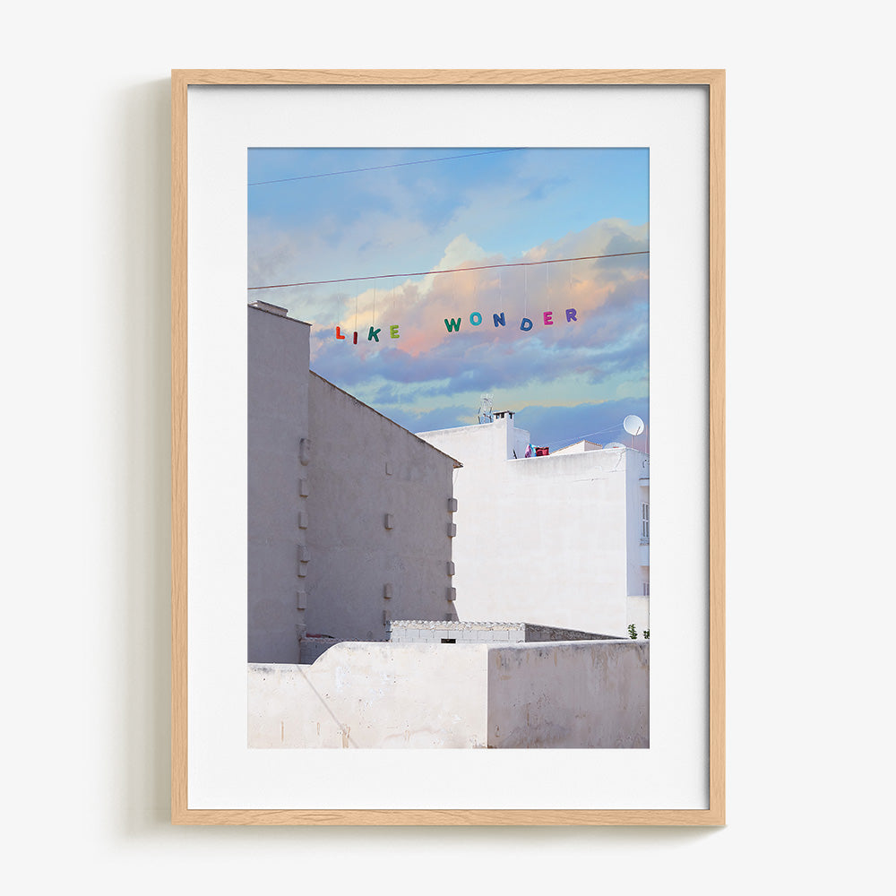 Like Wonder print by James Needham in a natural wood frame White buildings below a blue and pink cloudy sky with LIKE