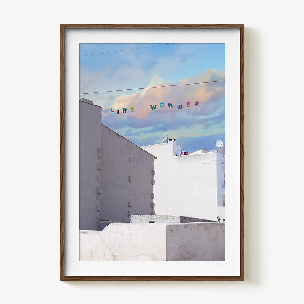 A print in a dark wood frame depicting white buildings under a blue sky with colorful letters spelling LIKE WONDER