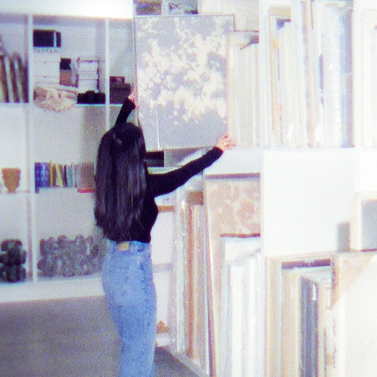 A person with long dark hair, wearing a black long-sleeve top and light blue jeans, stands in a room filled with shelves and framed art pieces, holding up a framed artwork.