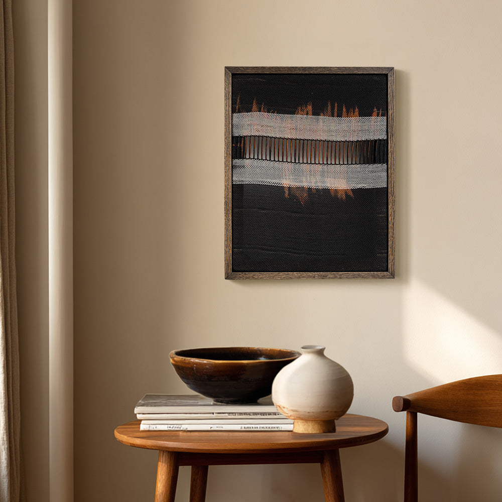 Dark textile art mounted in a wooden frame hanging on a wall above a small wooden table with pottery