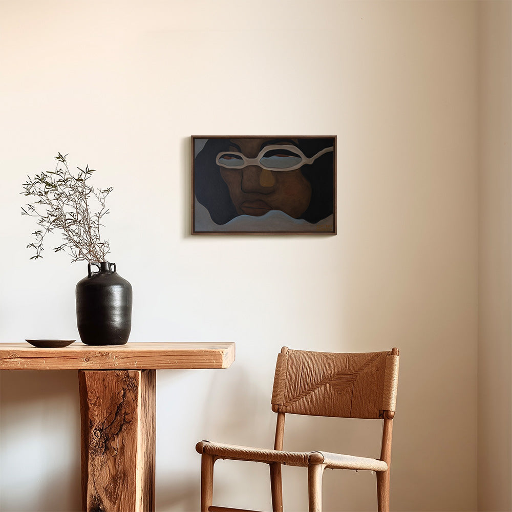 A framed painting of an abstract face wearing glasses hanging on a plain wall above a wooden table and chair