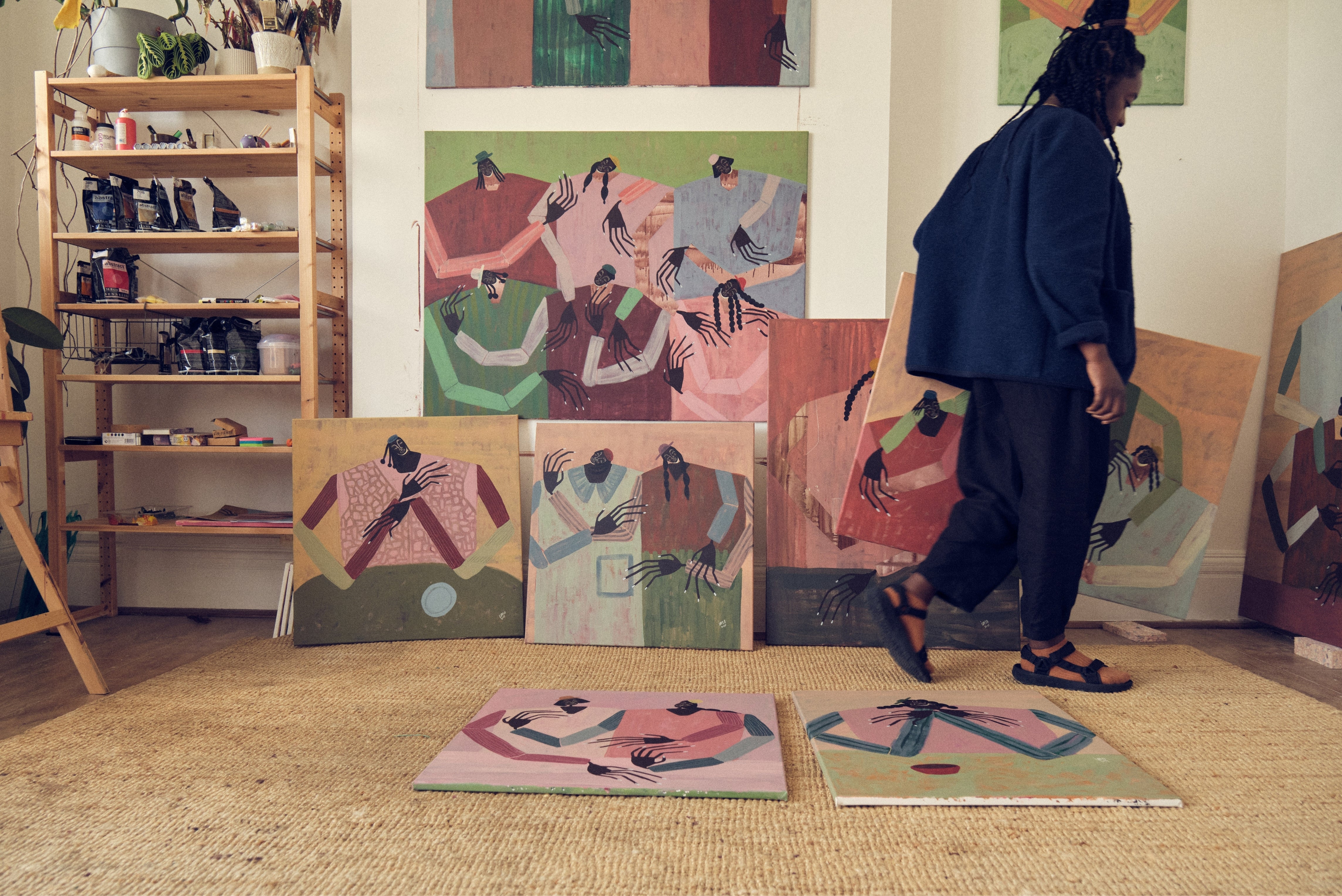 A person in a dark blue shirt and black pants walks through an art studio filled with colorful abstract paintings on the wall and floor. The room has a woven beige rug, a wooden shelving unit with art supplies, and natural light coming from a window.
