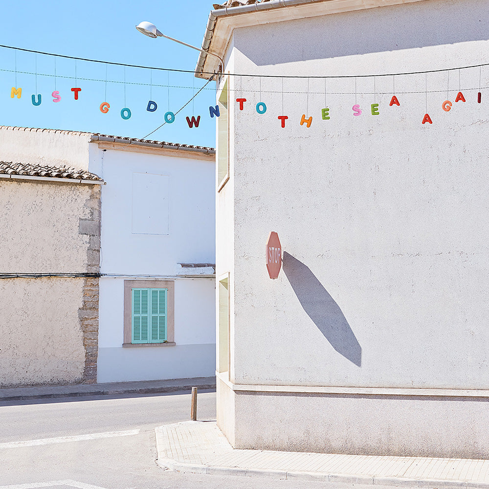 Down to the Sea print by James Needham Colorful letters strung across a street between white buildings without a frame