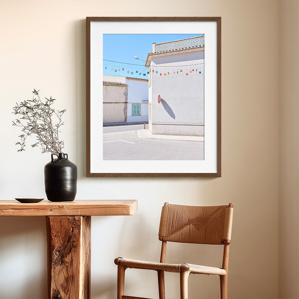 Coastal village photo print with white buildings colorful bunting and a sunlit street in a brown wood frame