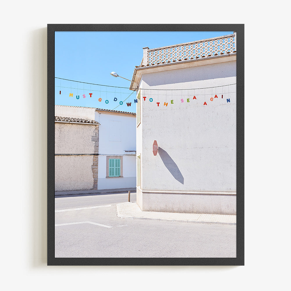 A framed print of a white building with a stop sign shadow and colorful letters spelling I MUST GO DOWN TO THE SEA AGAIN