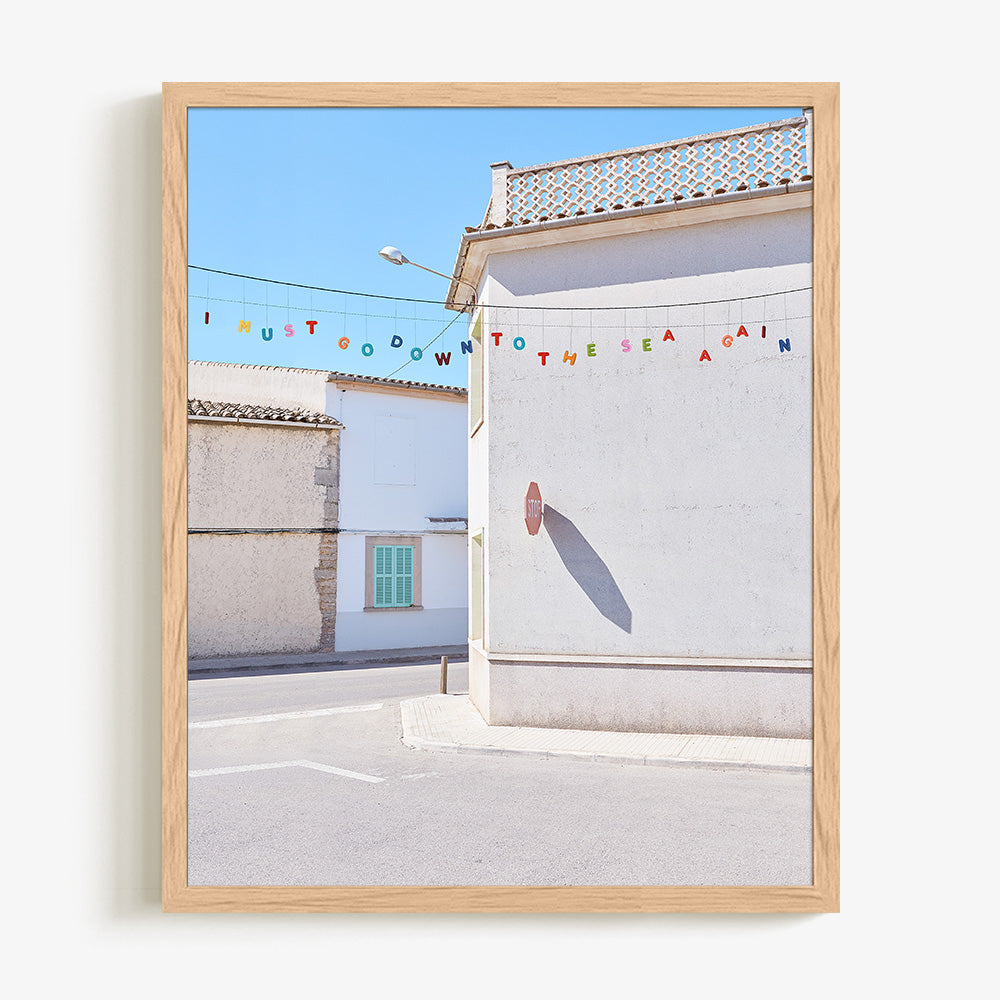 A light wood framed print of a white building with a stop sign shadow and colorful letters spelling I MUST GO DOWN TO