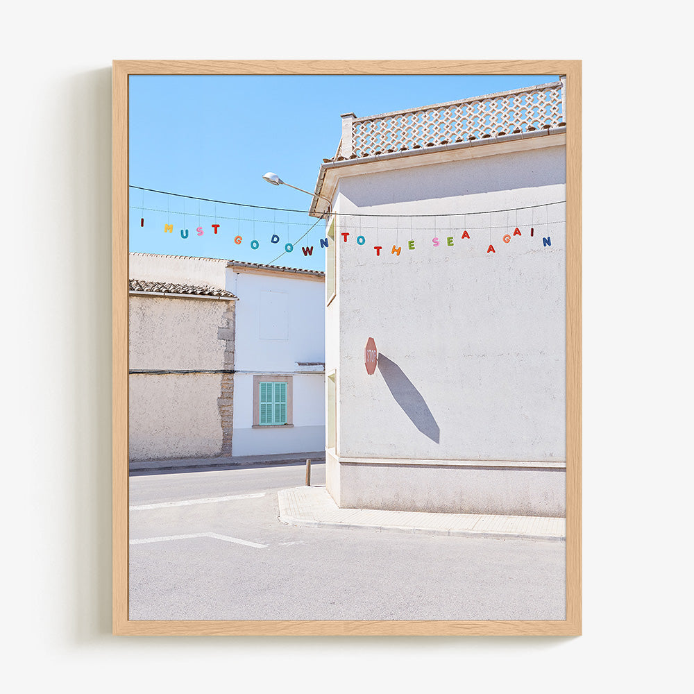 Coastal village photo print with white buildings colorful bunting and a sunlit street in a light wood frame