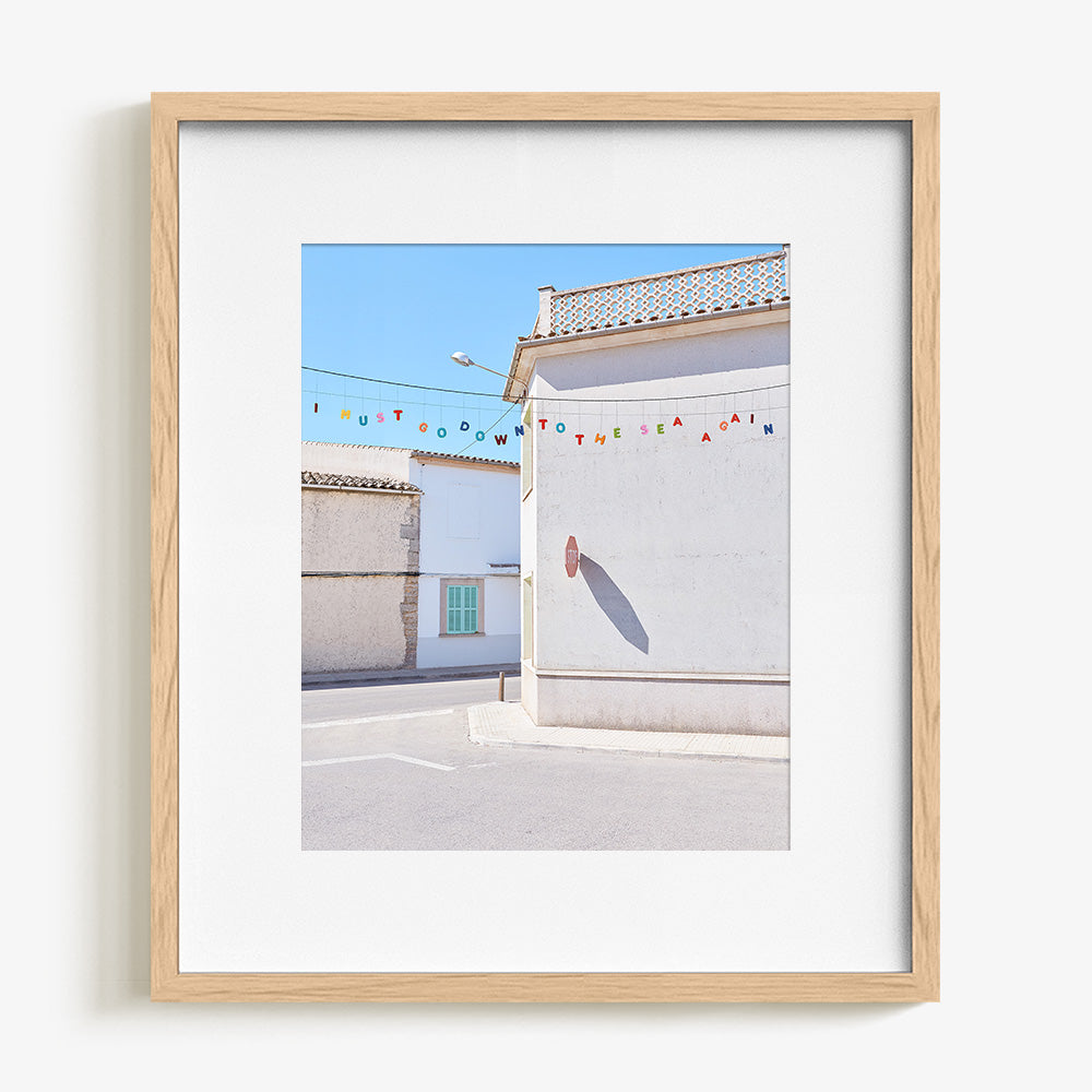 A light wood framed print with a white border showing a white building with a stop sign shadow and colorful letters