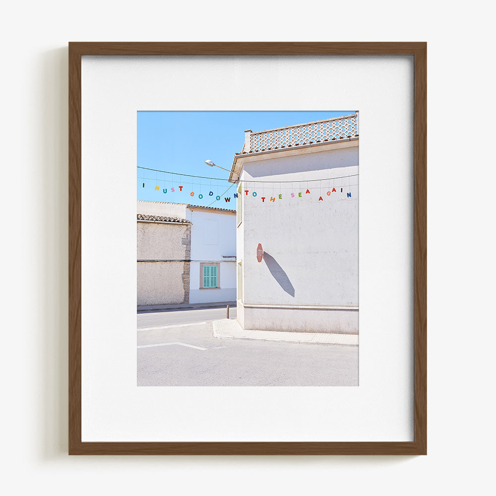 A dark wood framed print with a white border showing a white building with a stop sign shadow and colorful letters