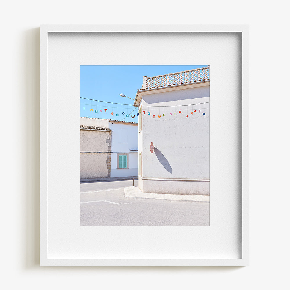 A white framed print with a white border showing a white building with a stop sign shadow and colorful letters spelling