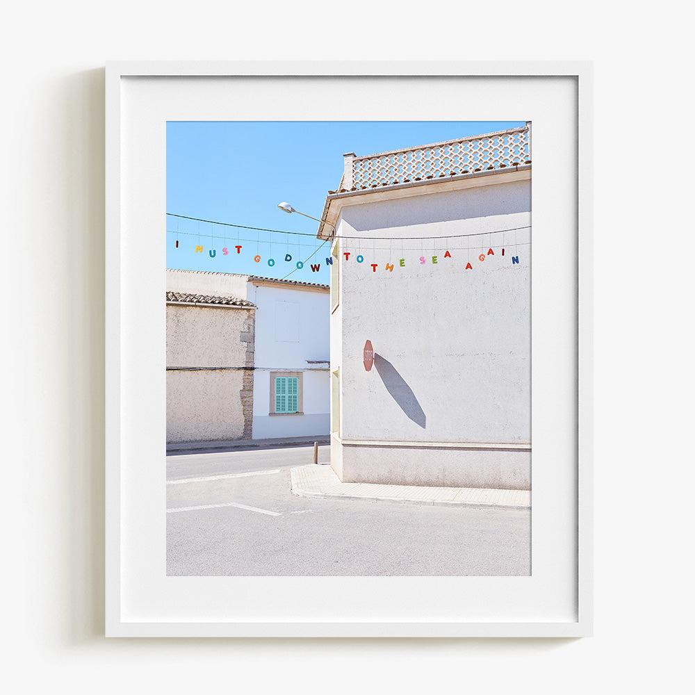Close-up vertical photo print featuring white stucco buildings with a green shutter and colorful bunting framed in white