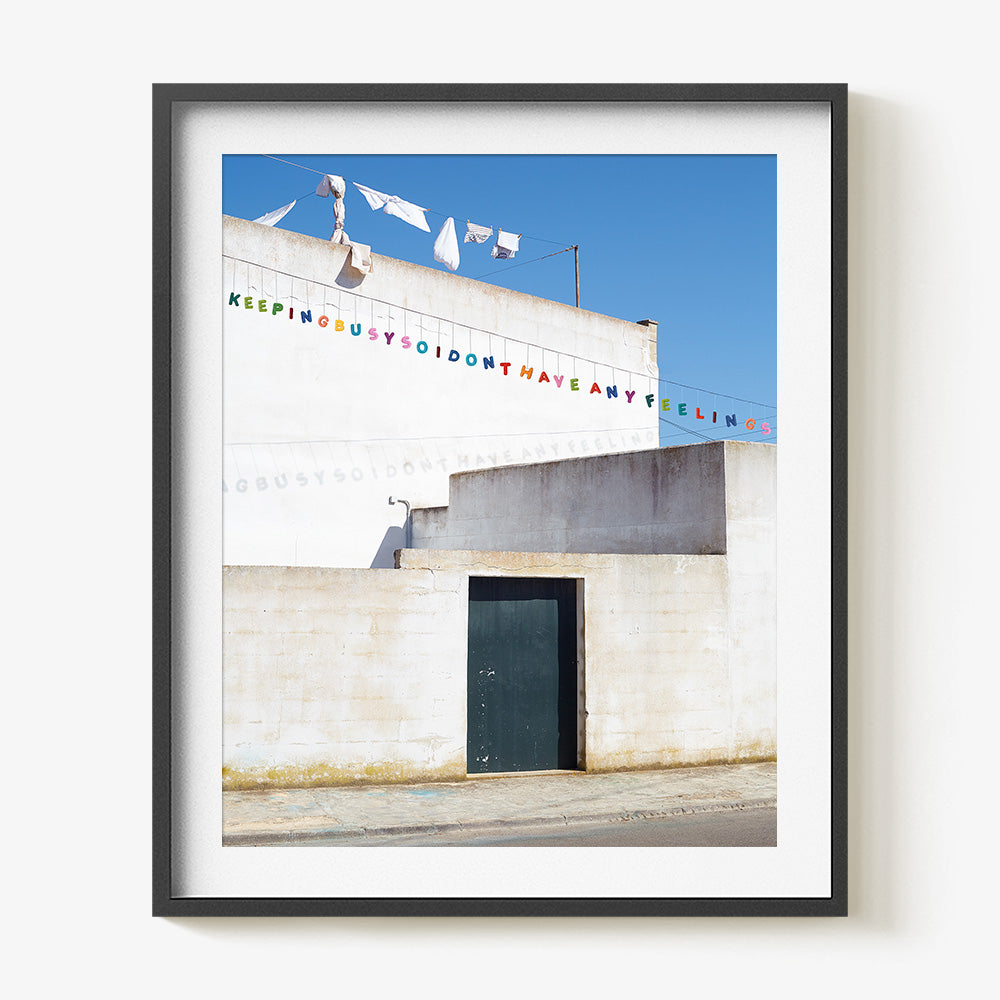 A white building wall with colorful text bunting mounted in a thin black frame