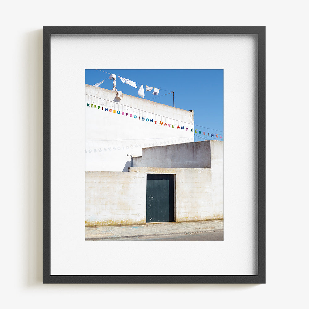 Framed print featuring a white building dark door and laundry line with colorful letters in a charcoal frame