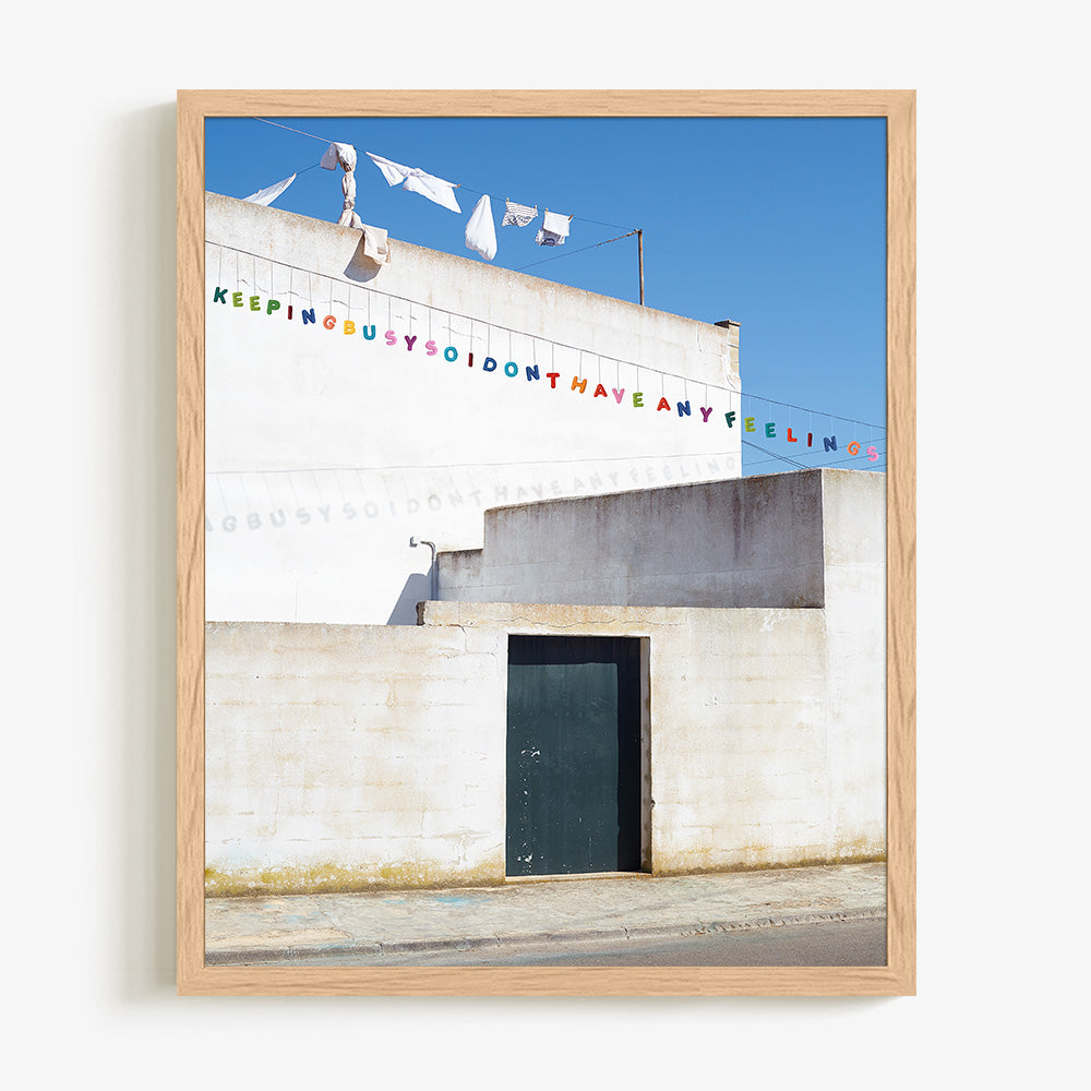 Print of a sunlit white building with hanging laundry and colorful banner text in a light wood frame