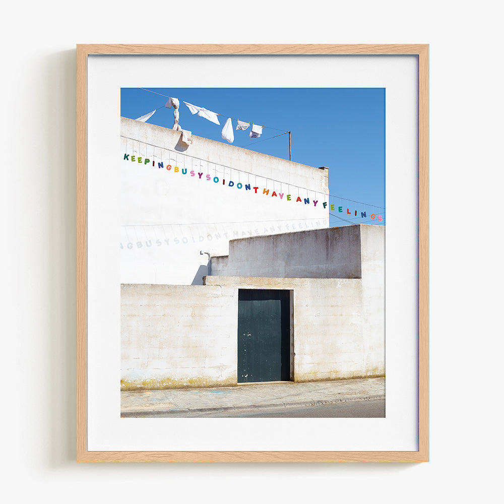 A framed print of a white building with a dark door and festive colorful text hanging across the sky