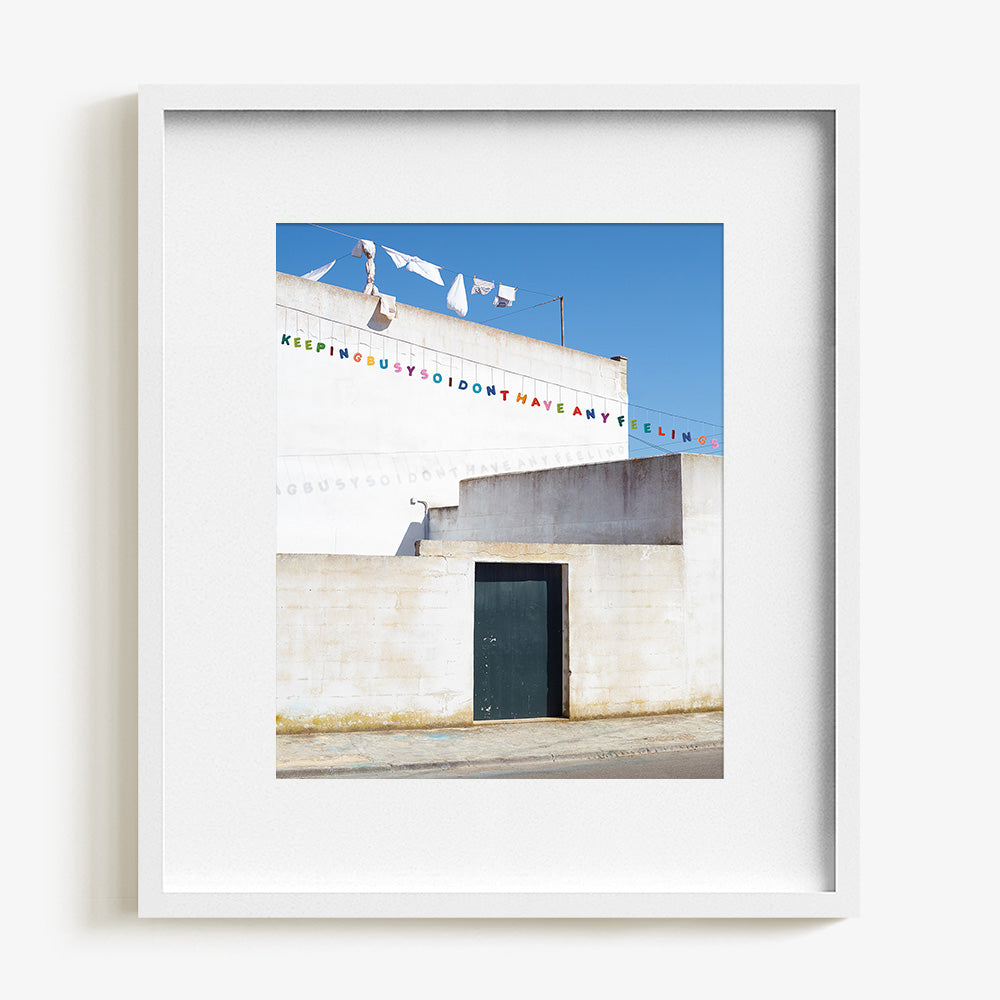 Print of a minimalist white architectural scene with hanging laundry and text banners in a thin white frame