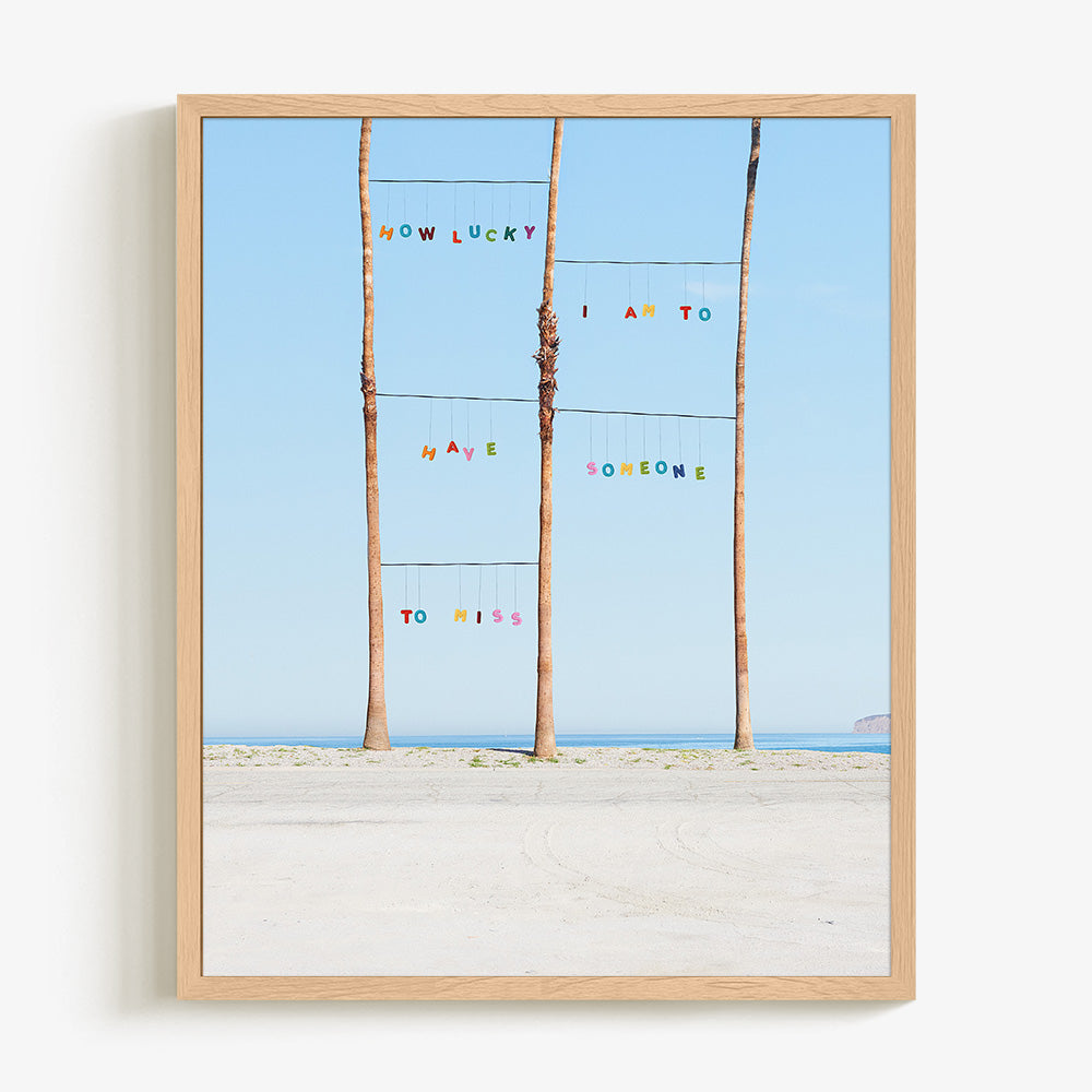Lucky print by James Needham Colorful letters suspended between palm trees on a beach in a light wooden frame