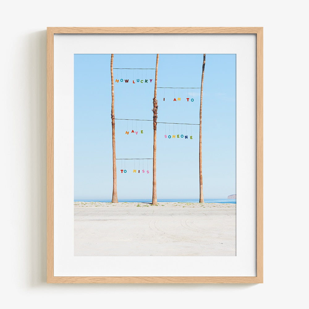Lucky print by James Needham Colorful letters suspended between palm trees on a beach in a light wooden frame with a