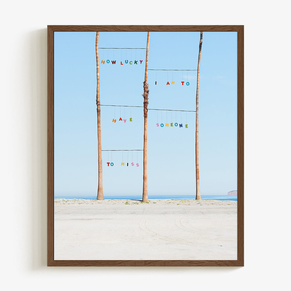 Lucky print by James Needham Colorful letters suspended between palm trees on a beach in a dark wooden frame