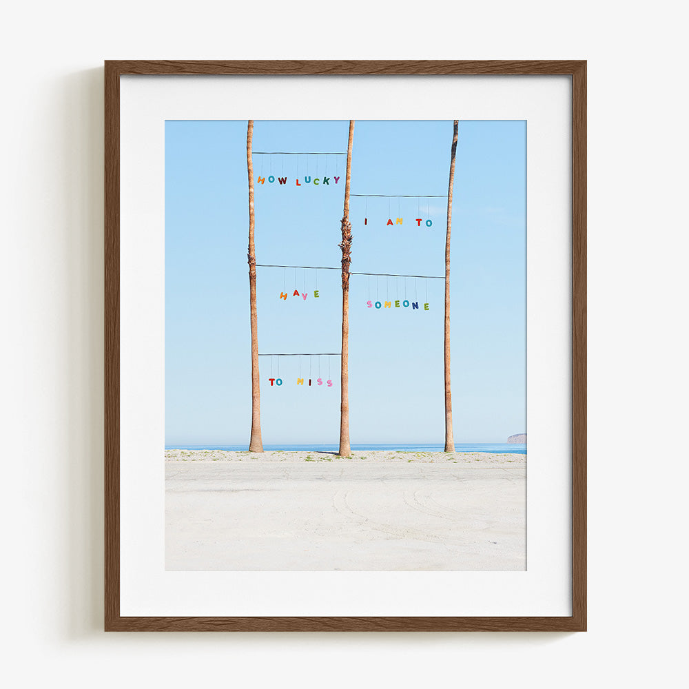 Lucky print by James Needham Colorful letters suspended between palm trees on a beach in a dark wooden frame with a