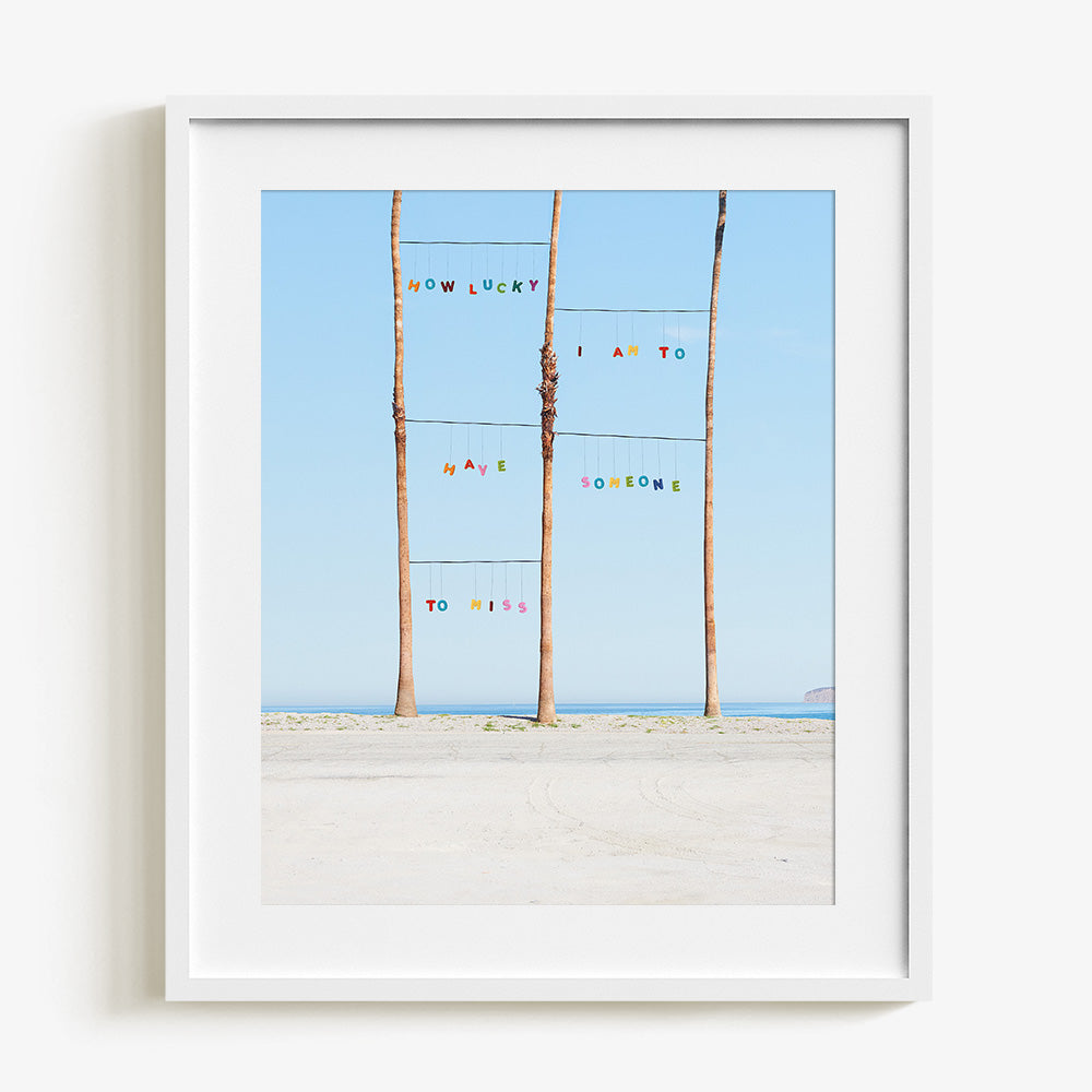 Lucky print by James Needham Colorful letters suspended between palm trees on a beach in a white frame with a white mat