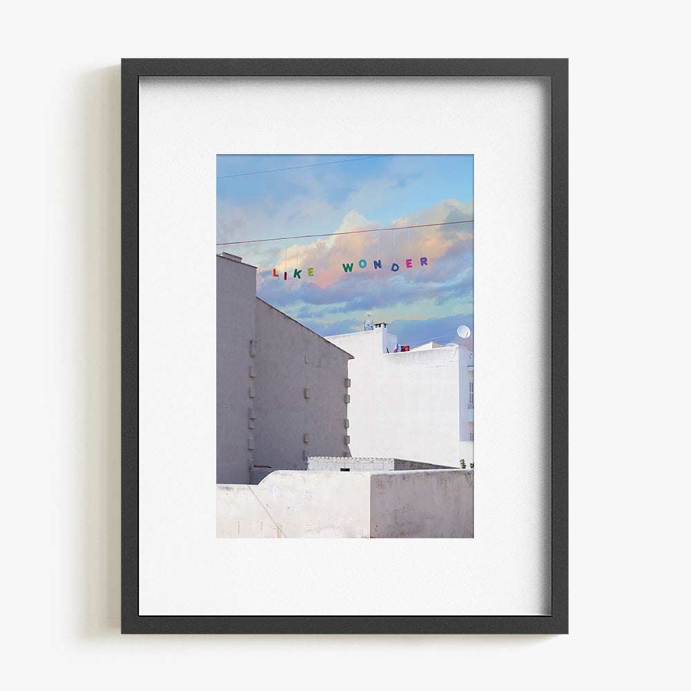 Art print of white buildings under a blue and purple cloudy sky with LIKE WONDER in colorful letters in a black frame