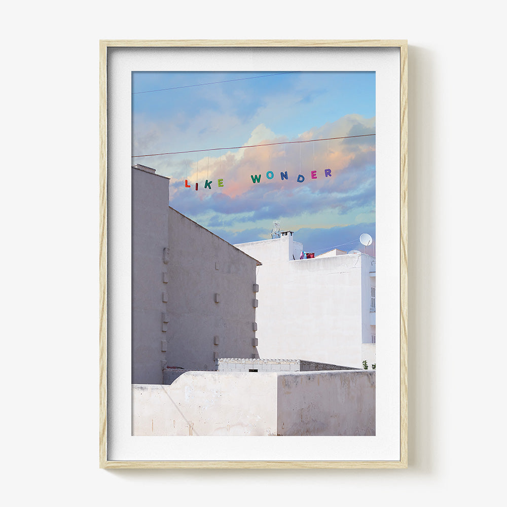 A print in a natural wood frame depicting white buildings under a blue sky with colorful letters spelling LIKE WONDER