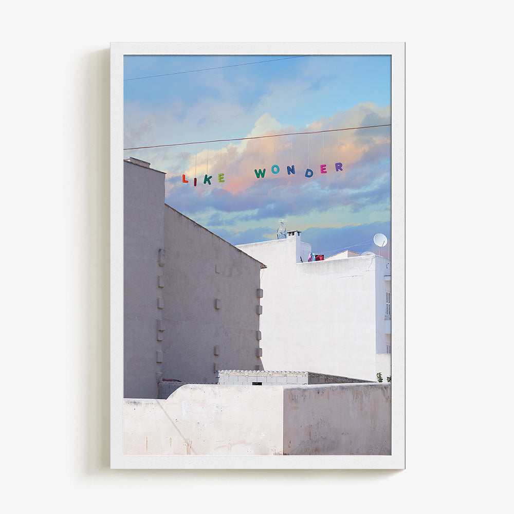 Art print of white buildings under a blue and purple cloudy sky with LIKE WONDER in colorful letters