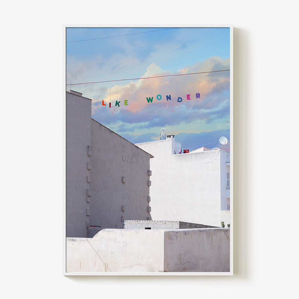 Art print of white buildings under a blue and purple cloudy sky with LIKE WONDER in colorful letters in a white frame