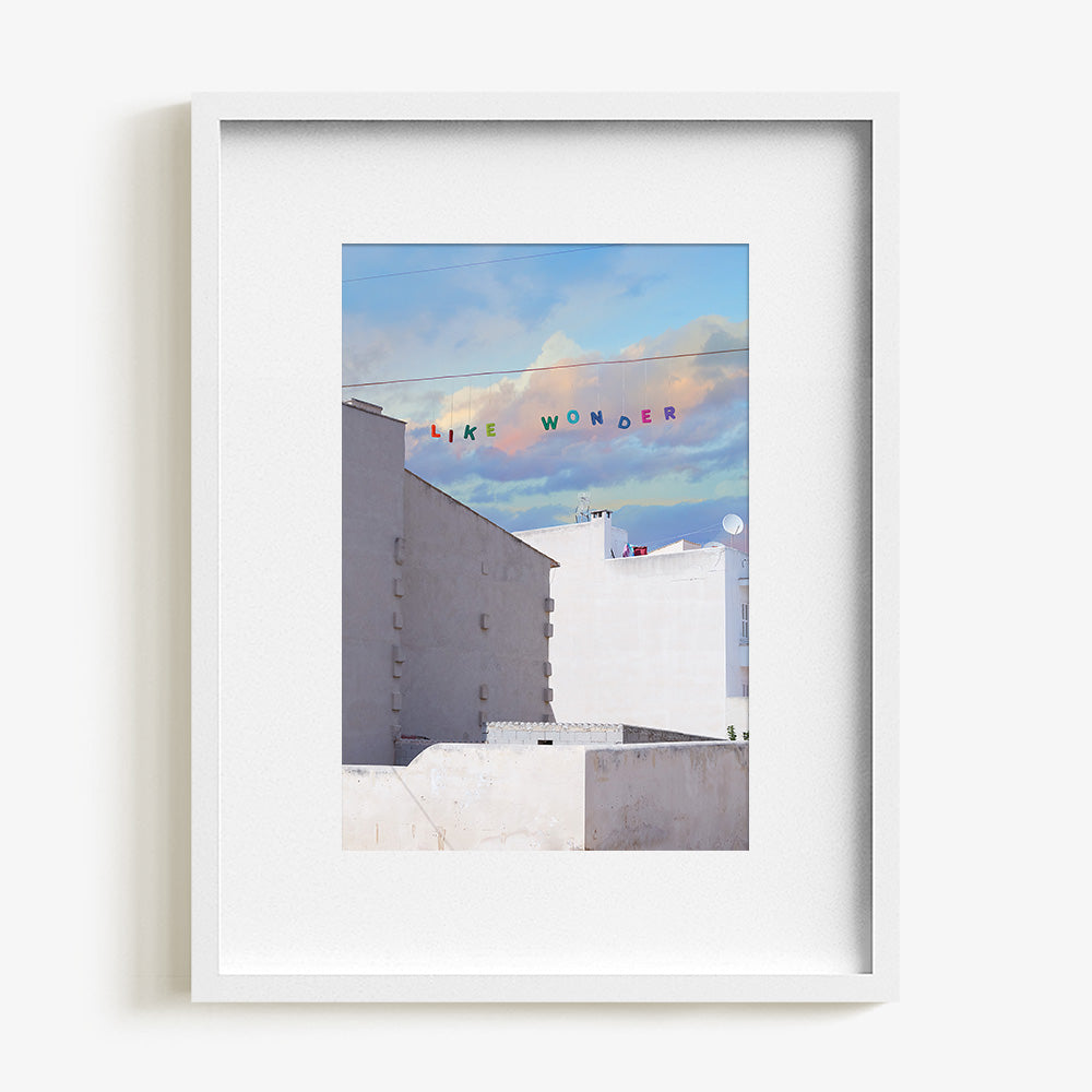 Like Wonder print by James Needham in a white frame White buildings below a blue and pink cloudy sky with LIKE WONDER