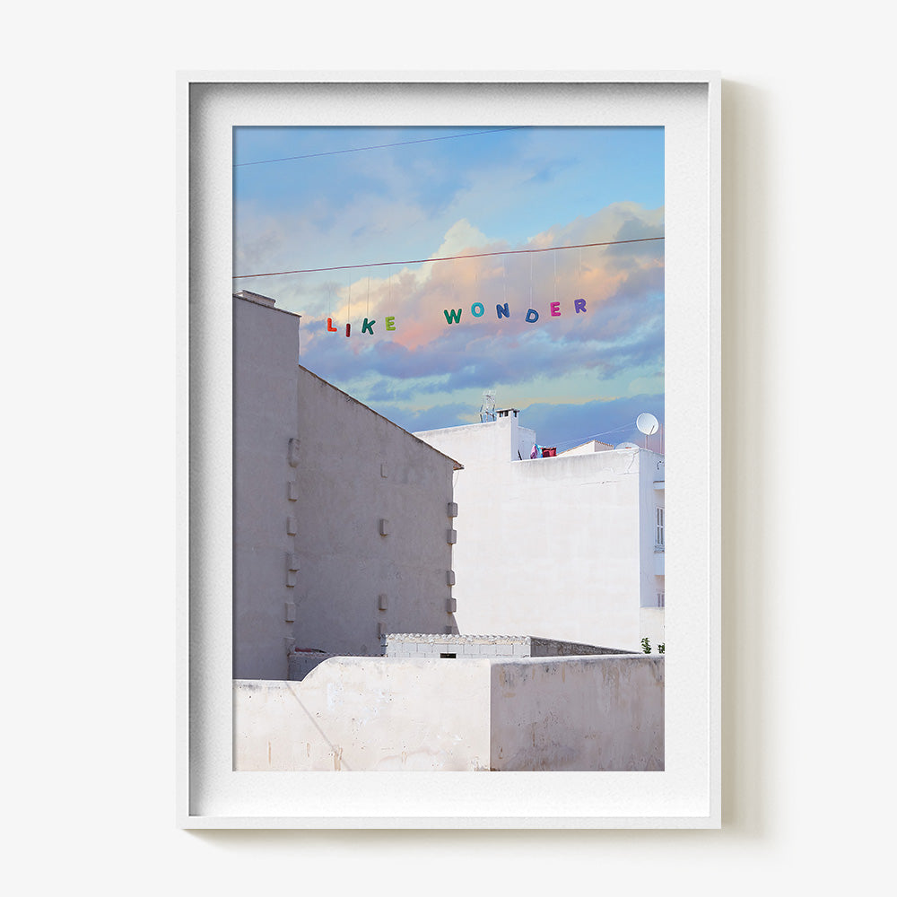 A print in a white frame depicting white buildings under a blue sky with colorful letters spelling LIKE WONDER strung