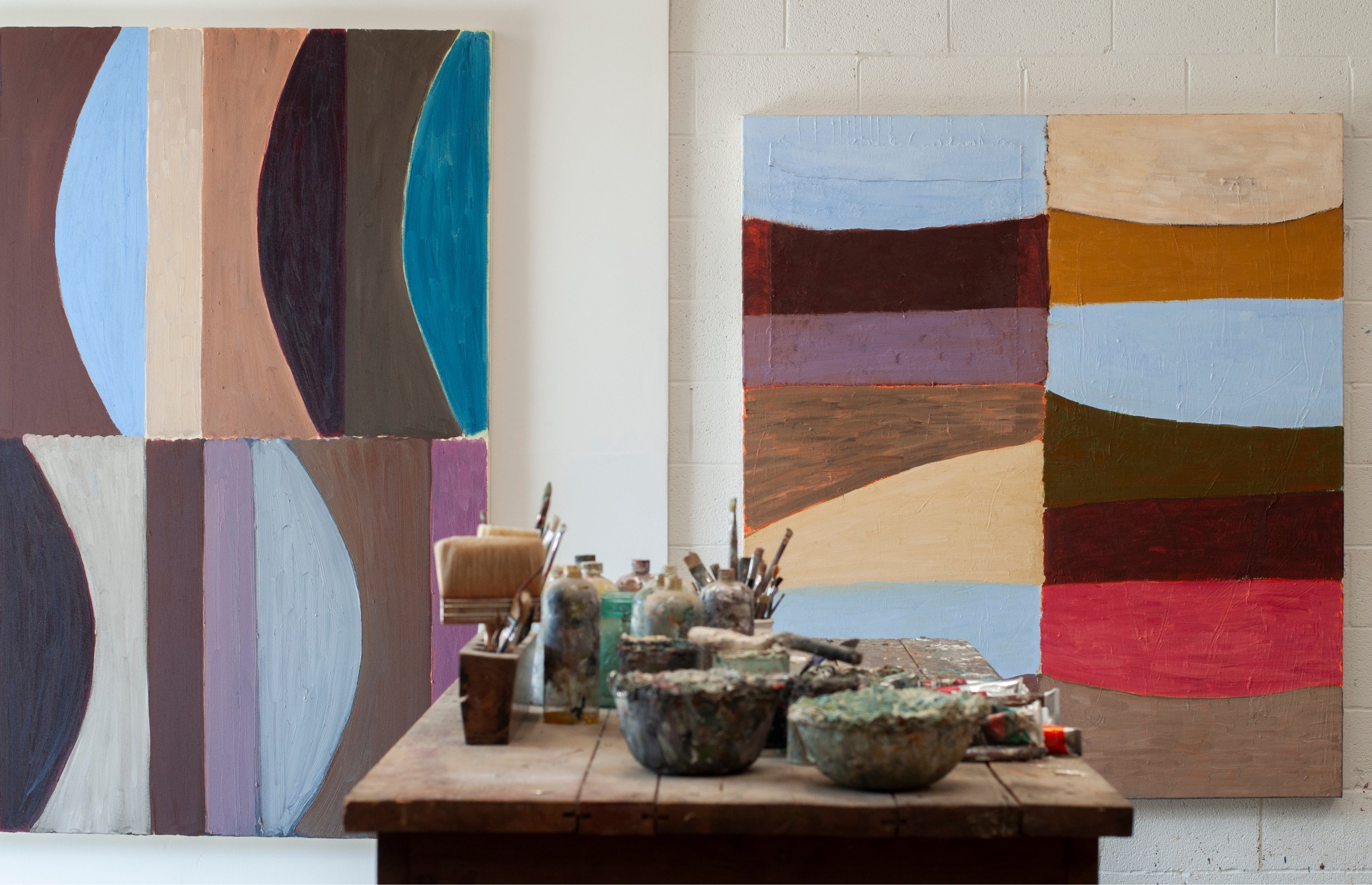 Two large abstract wall paintings with geometric color blocks hang on a white brick wall above a wooden worktable filled with paintbrushes, jars, and bowls in an artist studio.